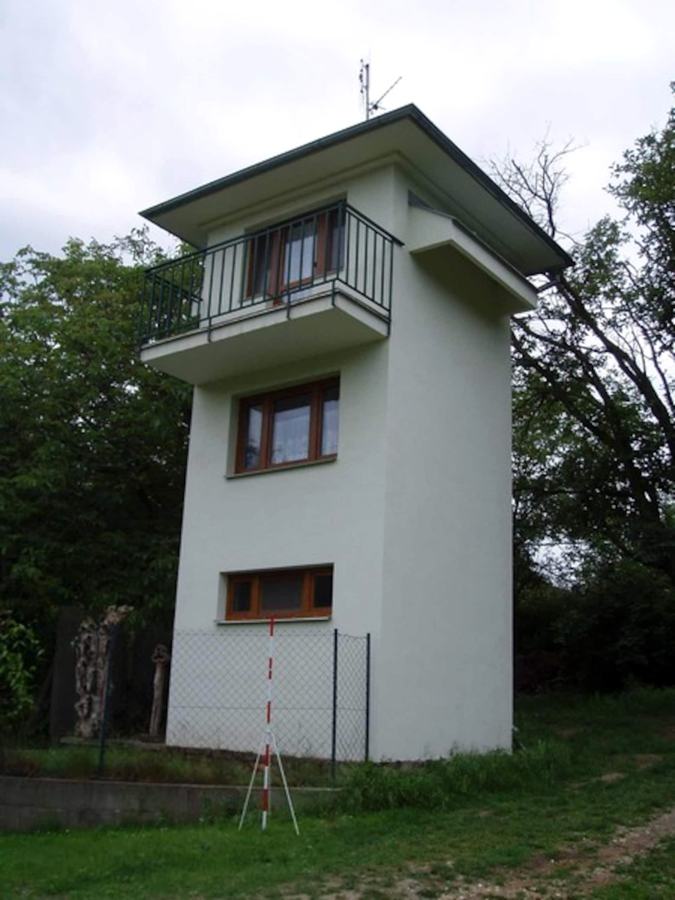 Bílý úzký třípatrový domek s balkonem nahoře, kolem zahrada